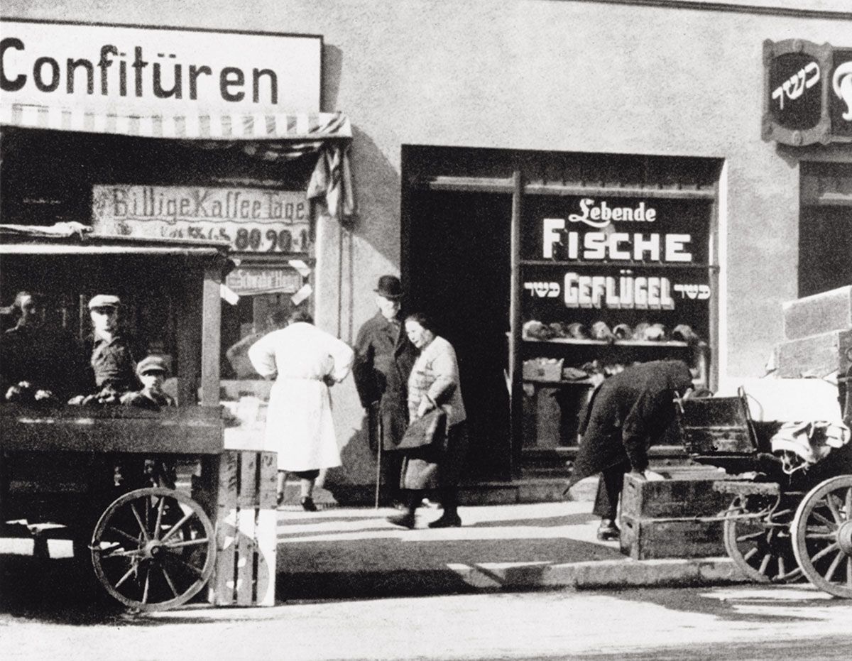Ulica z witryną sklepu z niemieckimi napisami i grupa osób przed nią.