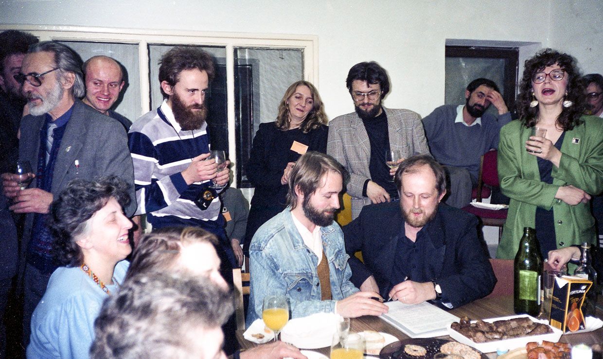 Группа людей за столом с едой и напитками. Двое мужчин на переднем плане, улыбаясь, подписывают документ
